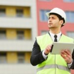 Encargado de obra con casco y tablet revisando un edificio durante la planificación constructiva.