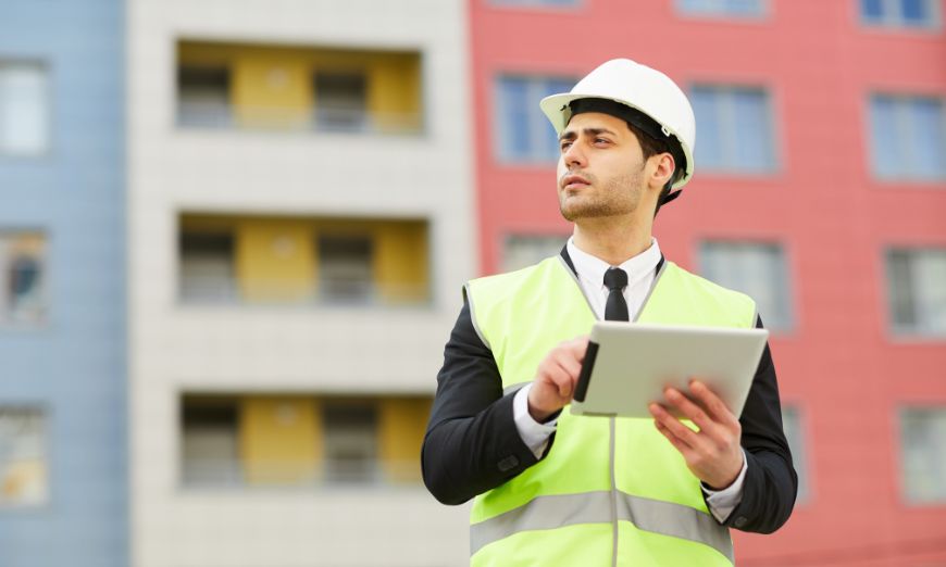 Encargado de obra con casco y tablet revisando un edificio durante la planificación constructiva.